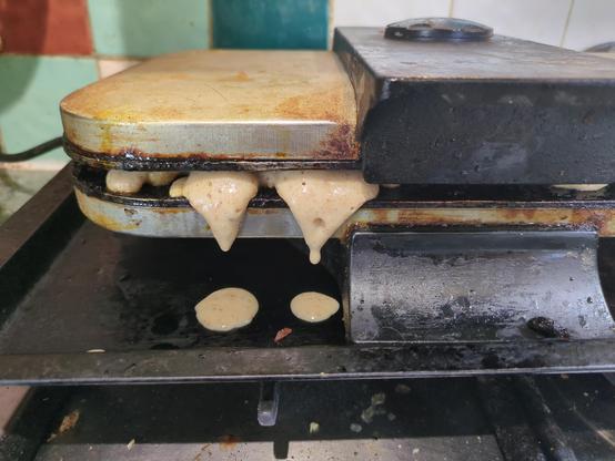 The left half of the front of an oldish wafflemaker. There are colourful square tiles on the wall bwehing it. Batter is oozing out and steam is rising.