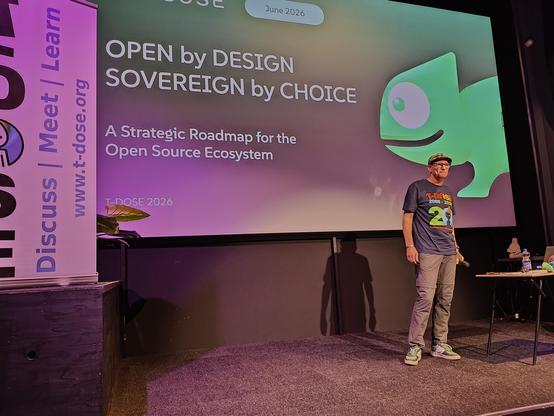 Emiel Brok on stage, a slide showing: "Open by DESIGN, SOVEREIGN by CHOICE -- A Strategic Roadmap for the Open Source Ecosystem" with a big stylished Geeko head.