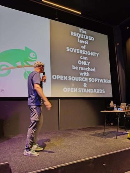 Emiel Brok on stage standing before a slide: left half a stylished green on white Geeko, on the right white on black: "The REQUIRED level of SOVEREIGNTY can ONLY be reached with OPEN SOURCE SOFTWARE & OPEN STANDARDS"
