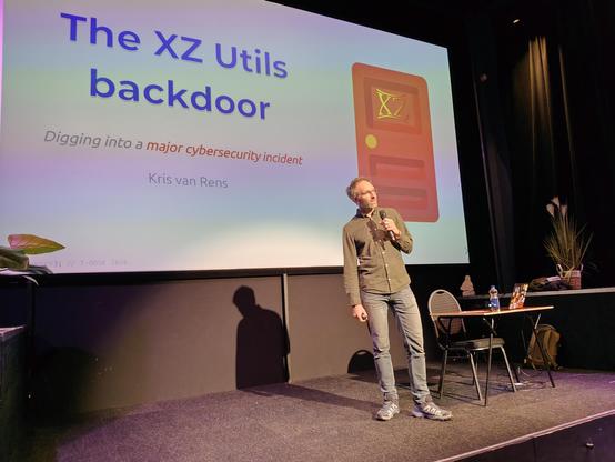 Kris van Rens  on stage.  Slide with blue on white letters: "The XZ Utils backdoor -- Digging into a major cybersecurity incident"