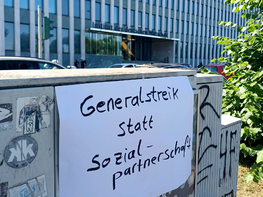 An einem Stromkasten hängt ein Plakat: "Generalstreik statt Sozialpartnerschaft"; im Hintergrund das "Institut der deutschen Wirtschaft" am Konrad-Adenauer-Ufer