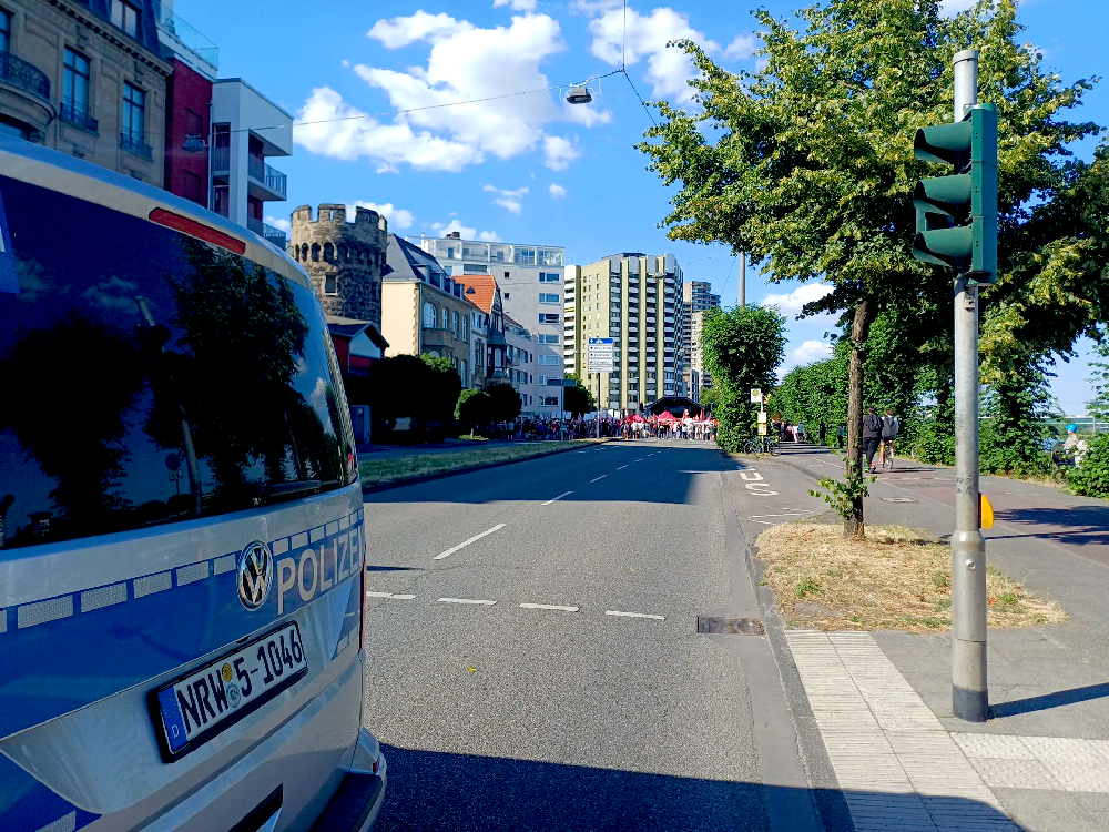 Ein Polizeiauto steht quer auf der von Häusern und Laubbäumen begrenzten Rheinuferstraße im Kölner Norden; weit hinten stehen Demonstrant*innen vor der Bühne des DGB