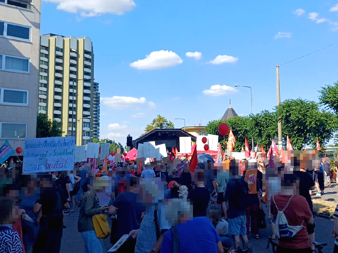 Kundgebung an der Kölner Bastei mit roten Fahnen  von Gewerkschaften und Parteien; im Hintergrund Hochhäuser und grüne Laubbäume.