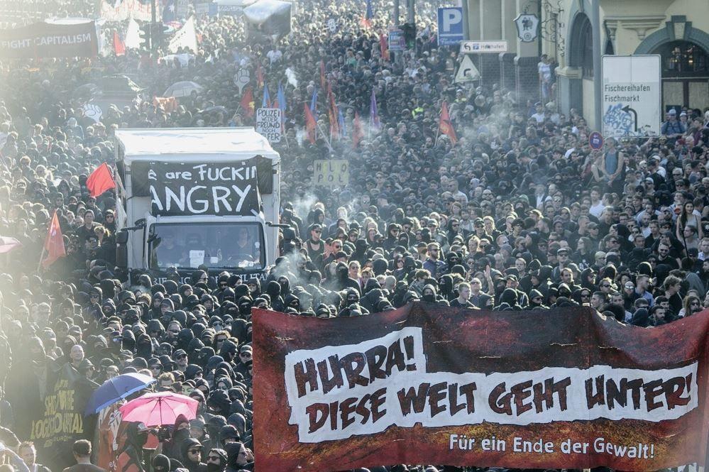 Großer Straßenprotest mit tausenden Teilnehmenden, vorn ein rotes Banner „Hurra! Diese Welt geht unter! Für ein Ende der Gewalt!“ und ein Lieferwagen mit dem Schriftzug „are FUCKIN ANGRY“.
