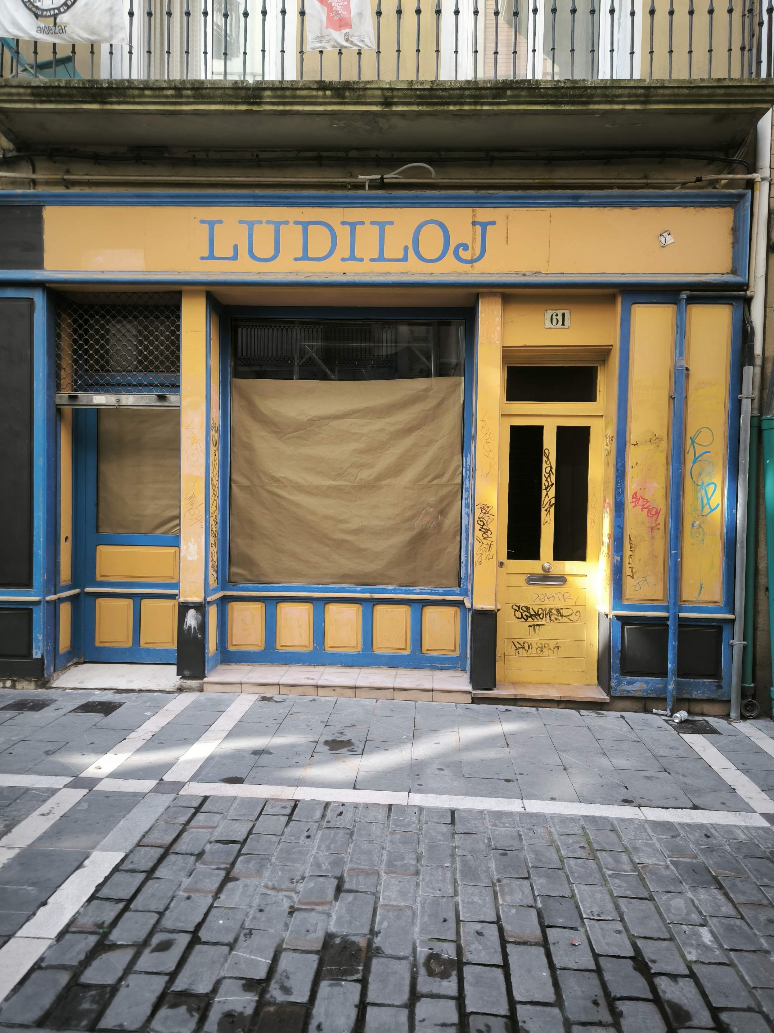 Una fachada de tienda antigua y desgastada, pintada principalmente en amarillo mostaza y azul vibrante. En la parte superior, un letrero amarillo con letras azules dice "LUDILOJ". La tienda parece cerrada o abandonada, con la mayoría de sus grandes ventanas cubiertas por papel marrón desde el interior. A la izquierda, una ventana más pequeña tiene una rejilla metálica. A la derecha, una puerta amarilla con el número "61" arriba está cubierta de grafitis coloridos y desgastados, al igual que los paneles inferiores de la fachada. La pintura de la tienda se ve descascarada y envejecida. El suelo frente a la tienda es un pavimento de adoquines grises y losas más claras con líneas blancas, que parece húmedo en algunas partes. Arriba, se ve un balcón con barandilla de hierro forjado.