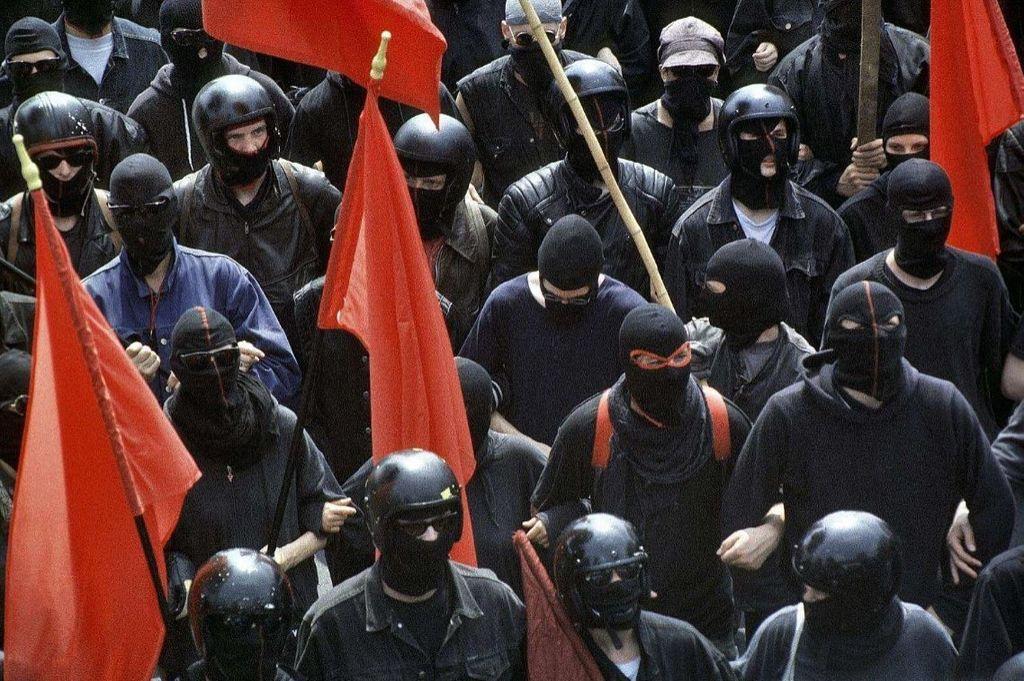 Vermummte Demonstrierende in schwarzer Kleidung und Schutzhelmen ziehen in dichter Formation durch die Straße und halten mehrere rote Fahnen.