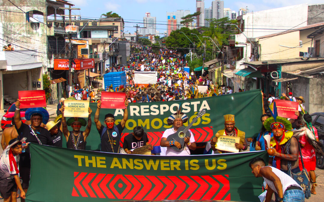 Image of an indigenous protest in an urban area of Brazil. The indigenous people in the foreground are holding a banner that reads: 
The answer is us
Source: https://apiboficial.org/2025/11/27/avaliacao-da-articulacao-dos-povos-indigenas-do-brasil-apib-sobre-a-cop30/
