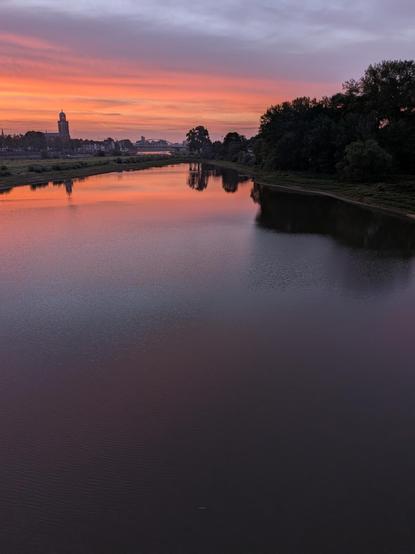 Uitzicht op nevengeul van de IJssel vanaf de spoorbrug bij Deventer. De lucht is bewolkt en wordt verlicht door de opkomende zon, waardoor het een kleurenpalet krijgt met roze, oranje lila. 
Op de voorgrond de spiegelgladde nevengeul. Op de achtergrond het silhouet van Deventer