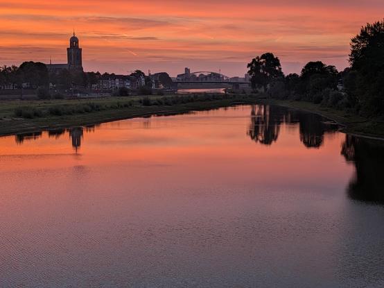 Uitzicht op nevengeul van de IJssel vanaf de spoorbrug bij Deventer. De lucht is bewolkt en wordt verlicht door de opkomende zon, waardoor het een kleurenpalet krijgt met roze, oranje lila. 
Op de voorgrond de spiegelgladde nevengeul. Op de achtergrond het silhouet van Deventer